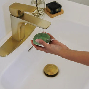 Hand holding a green Konjac sponge under running water in a sink.