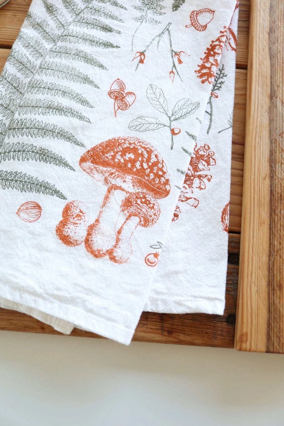 Close up of a white tea towel with fern fronds, mushrooms, nuts, and berries.