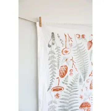 Close up of a white tea towel with fern fronds, mushrooms, and pine cones.