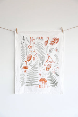 White tea towel with fern fronds, mushrooms, and pine cones.