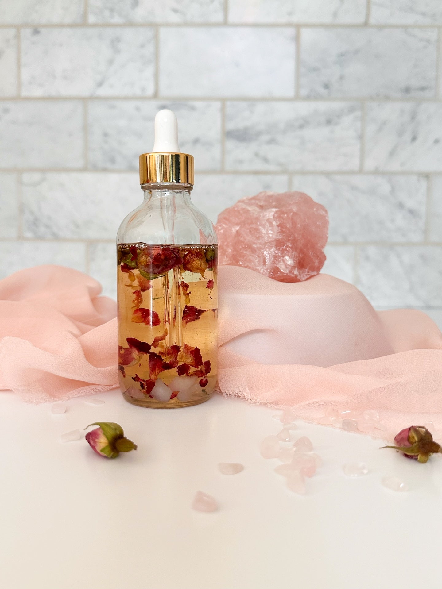 Rose petal and Rose Quartz body oil infusion with rose buds and Rose Quartz laying nearby.