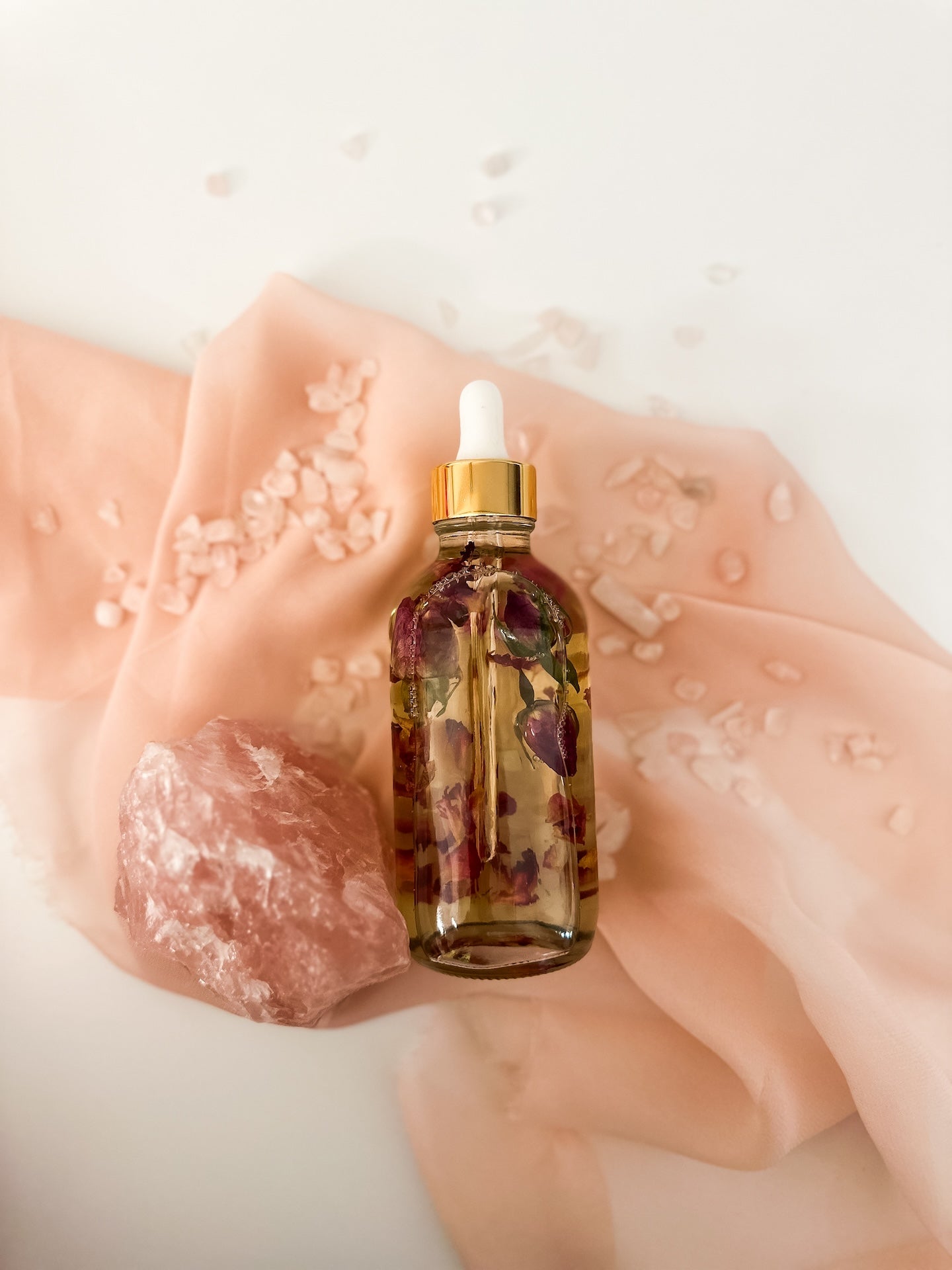 Rose and rose quartz body oil infusion laying down near rose quartz crystals and pink sheer fabric.