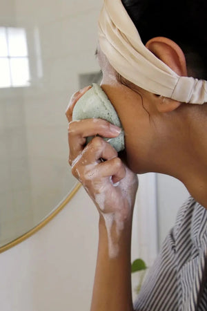 Woman using a green Konjac sponge on her face. Suds are running down her hand.